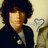 Pixelated image of a person with dark curly hair wearing a dark jacket over light shirt, holding a blue question mark symbol against a beige background.