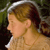 Pixelated profile view of a person with light brown hair tied back, wearing a light-colored top against a dark green and brown background.