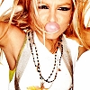 A person with long light brown hair wearing a white top and multiple beaded necklaces, with a bubblegum bubble in their mouth.