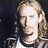 A pixelated image of a person with long light brown hair and a goatee, wearing a dark jacket over a white shirt against a black background.