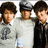 Three pixelated figures stand side-by-side; left figure wears light gray shirt with dark emblem, middle figure wears white shirt and dark cap, right figure wears dark suit with light shirt.