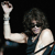 Pixelated figure with dark curly hair wearing dark sleeveless top, flexing right arm with clenched fist raised high against black background.
