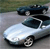 Two cars on a road, one silver in the foreground and one dark blue in the background, both with visible wheels and windows.