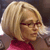 Pixelated close-up of a person with short blonde hair wearing red eyewear and a red top against a dark background.