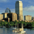 Pixelated cityscape with tall buildings, greenery, and water reflection under a blue sky.
