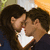 Two figures with dark curly hair embrace closely, one figure's face partially visible in profile while the other is turned toward them; a white heart shape appears between their faces against a blurred background.