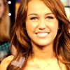 A close-up of a person with long brown hair wearing a light-colored top and a delicate necklace, smiling with visible teeth against a blurred background.