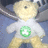 Pixelated static image of a light-colored teddy bear wearing a white shirt with a green circular emblem featuring a white cross-like symbol on its chest.