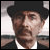 Static pixelated image of a person wearing a dark hat and dark jacket over a white shirt, with a blurred background showing light and dark horizontal bands.