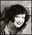 Static grayscale image of a person with dark hair styled in voluminous waves, wearing light-colored clothing against a neutral background.
