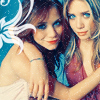 Two women pose closely together against a blue background with white floral patterns; one wears a red top and blue bracelet, the other a red dress.