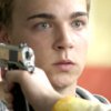 A person holds a silver handgun near their temple against a blurred background with orange and yellow tones.