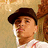 Pixelated image of a person wearing dark cap and white shirt with red stripes, standing against textured brown background.