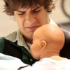 A person with brown curly hair wearing a dark green jacket over a white collared shirt holds a bald doll with a smooth light skin tone.