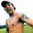 A pixelated image of a shirtless man with tattoos on his arms, standing outdoors with a grassy field and sky background, overlaid with red pixelated text.