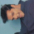 Pixelated image of a person with dark hair lying on a light blue surface, wearing a dark blue garment, with closed eyes and a neutral expression.