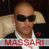 A person with a bald head wearing dark sunglasses, a white collared shirt, and a black jacket with a red emblem on the lapel, seated in a light-colored chair.