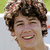 Pixelated close-up of a person with curly brown hair, closed eyes, and an open mouth showing teeth against a light background.