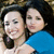 Two people with dark hair pose closely together, one wearing a blue top, against a dark background with light-colored elements.