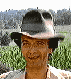 A person wearing a gray fedora with a black band, standing in a green field with trees in the background, smiling at the camera.