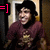 Static pixelated image of a person with dark hair wearing maroon clothing, standing in an interior space with light-colored trim and brown walls.