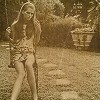 A sepia-toned static image shows a woman in a short patterned dress standing outdoors with one arm extended toward a stone structure.