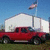 A red pickup truck with a white canopy is parked on grass under a blue sky, with a flagpole and flag visible in the background.