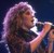 Pixelated image of a person with long light-colored hair holding a microphone against a dark background.
