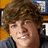 Pixelated close-up of a person with brown curly hair wearing a dark jacket over a light shirt, set against a blurred background with orange and blue tones.