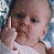 A pixelated image of a baby with light skin and brown hair wearing a white top, holding two fingers up near the face against a blue and white background.