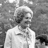 Pixelated grayscale image of a person in a white outfit with a light-colored head covering, standing against a textured background.