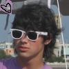 A person with dark curly hair wearing white-framed sunglasses and a purple shirt, with a pixelated heart symbol in the top-left corner.