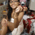 A woman in a red and white outfit poses with one hand near her face, holding a small object, against a blurred background.