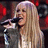 A person with long blonde hair sings into a microphone, wearing light-colored clothing against a dark background with red accents.