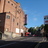 Pixelated image of a light brown building with multiple windows and a dark base, set against a blue sky with greenery and a red vehicle in the background.